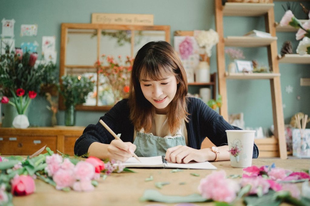 A young Asian woman florist writing notes in a vibrant floral workshop, smiling.